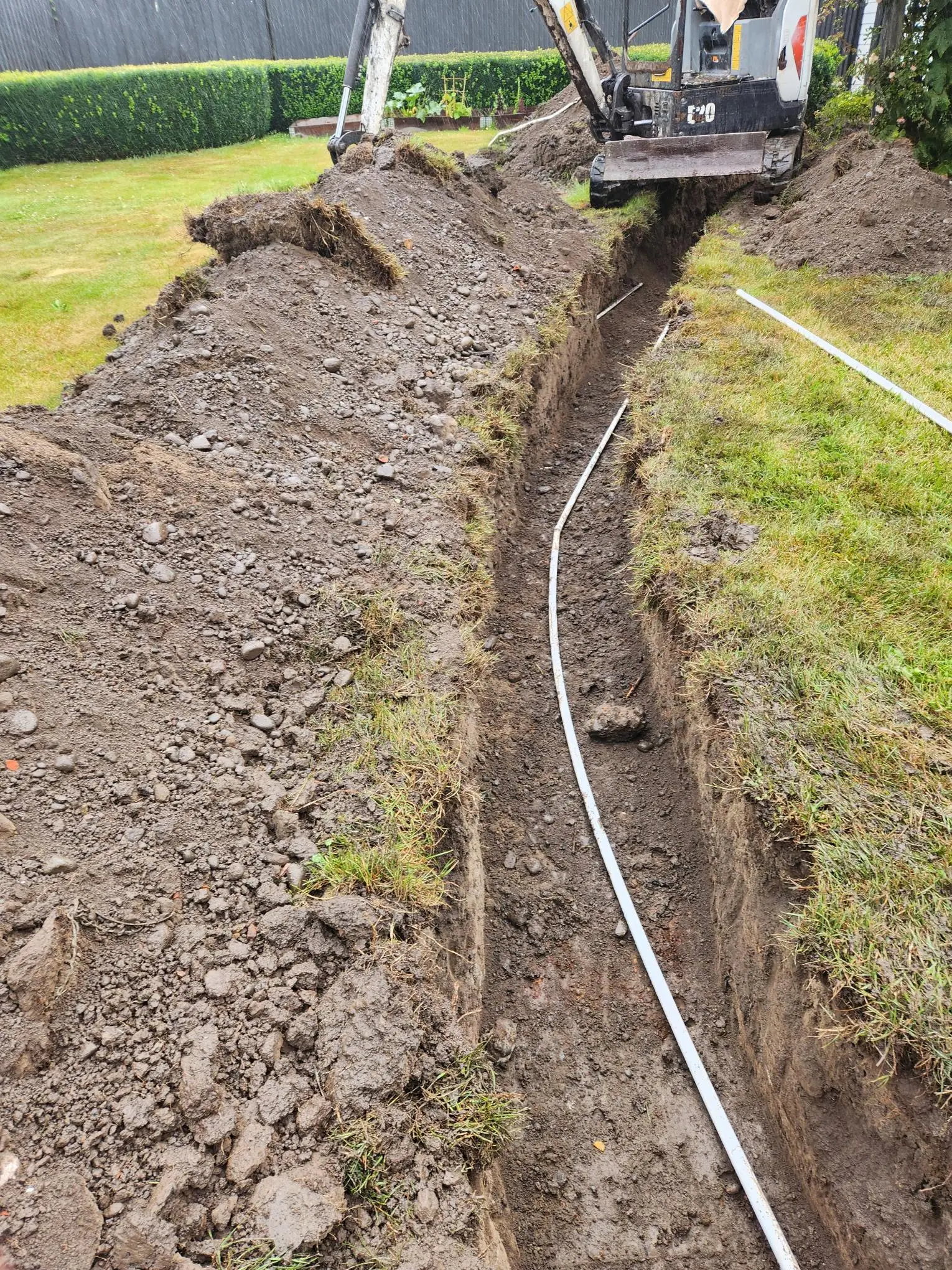 Mini digger digging a narrow trench with drainage pipe laid beside it